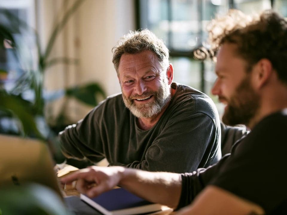 A man in his fifties smiles as a younger colleague points at a laptop beside a blue notebook