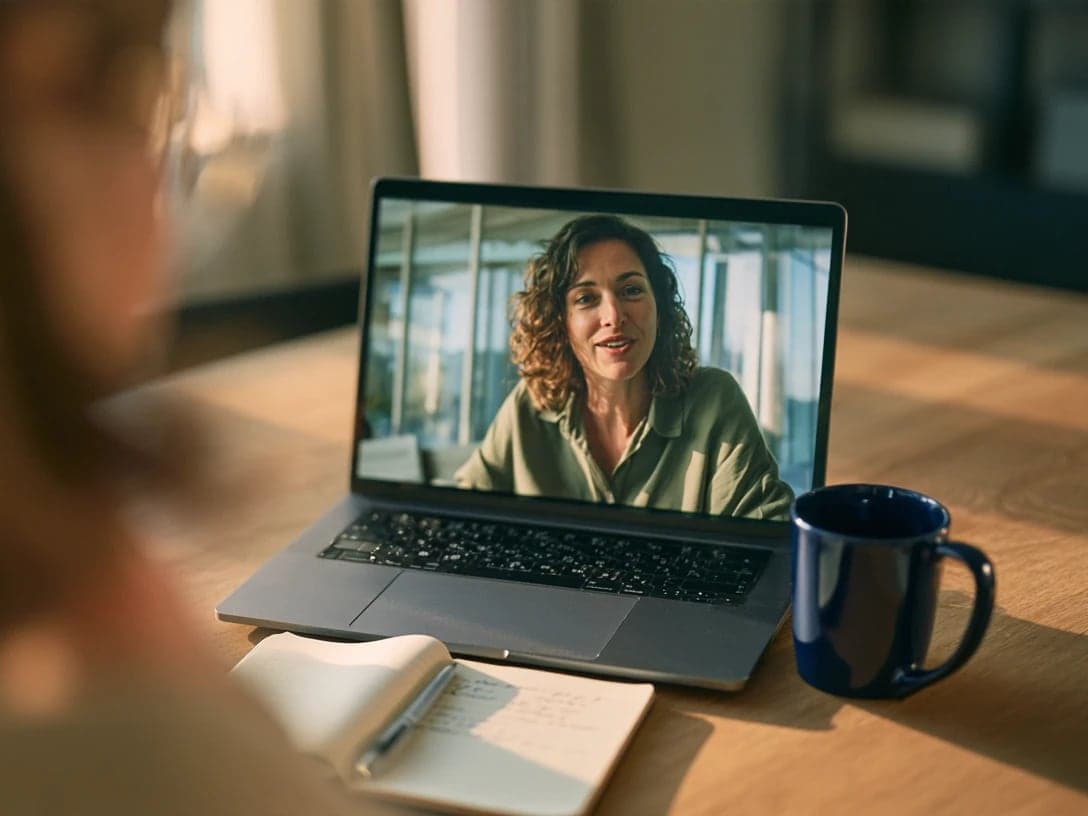 Someone on a laptop video call with a smiling woman, next to an open notebook and a blue mug