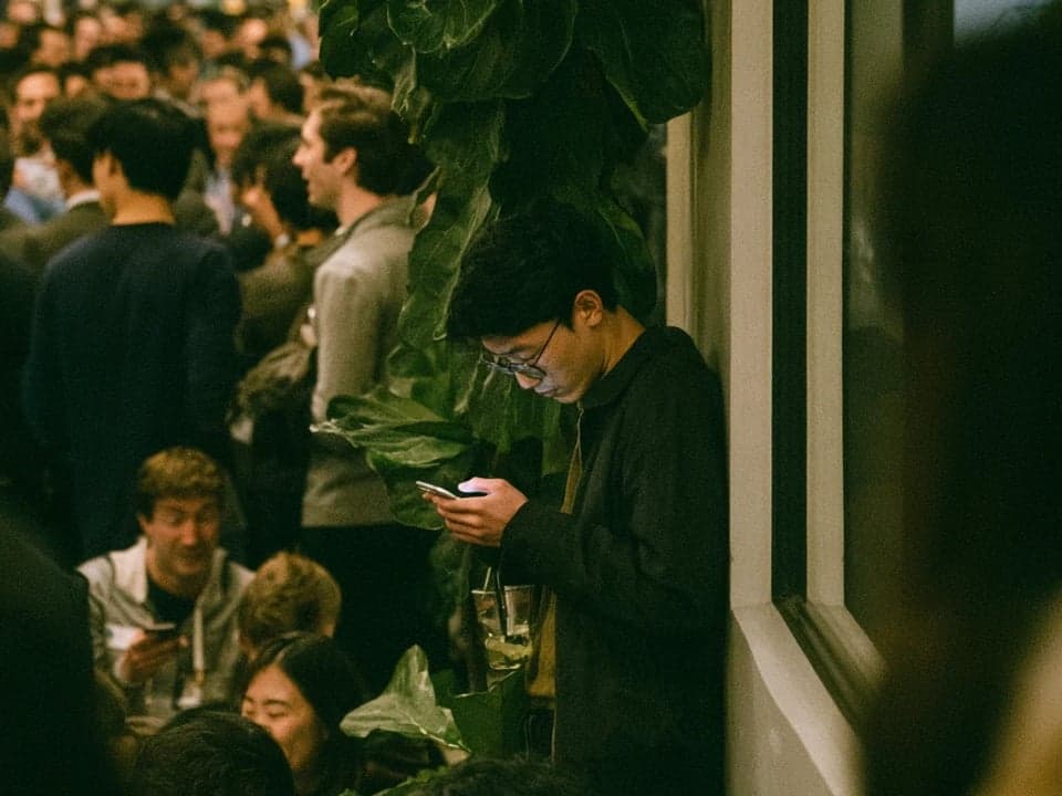 A young man in glasses leans against a wall, looking at his phone, while a crowded networking event goes on beside him