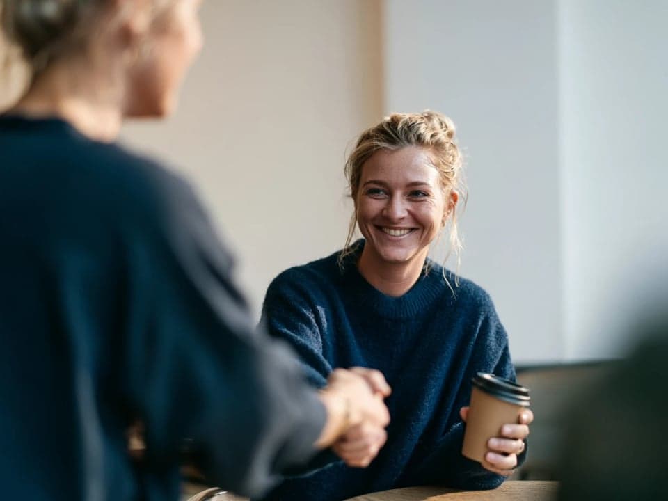 Two women shake hands across a café table, one smiling and holding a takeaway coffee